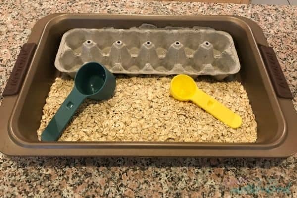 A sensory bin comprised of a baking pan containing the bottom half of an egg carton, oats, and measuring spoons.