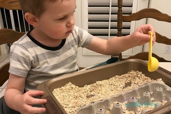 Child at a table with an easy sensory bin comprised of a metal baking pan, the bottom half of an egg carton, oats, and plastic measuring utensils, one of the fun and easy indoor preschool activities in the list.