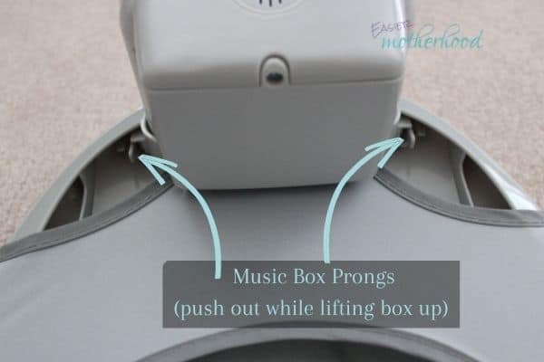 The back side of the ingenuity smartbounce automatic bouncer, with arrows detailing where the prongs are to open, with text conveying that pushing the prongs open while lifting the box up is how to remove it- a necessary step to remove the ingenuity bouncer cover.
