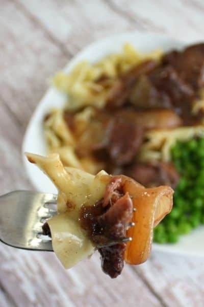 Beef onion wine sauce over pasta on a white plate, image has a fork with a bite in the foreground