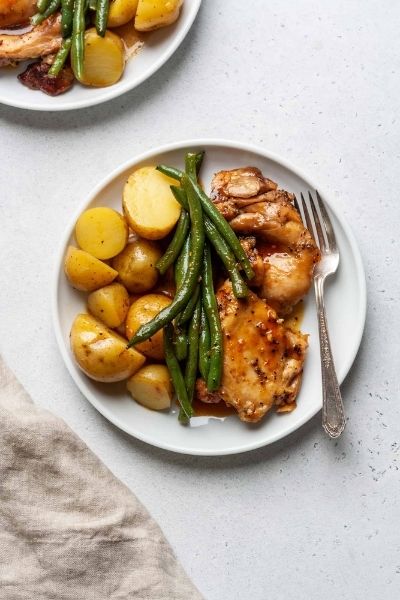 Chicken, potatoes, and green beans on a white plate with a fork. Second plate and napkin partially in the frame