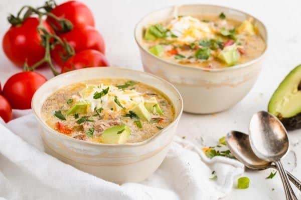 Two white bowls filled with slow cooker keto taco soup, with tomatoes, an avocado, and kitchenwares in the background