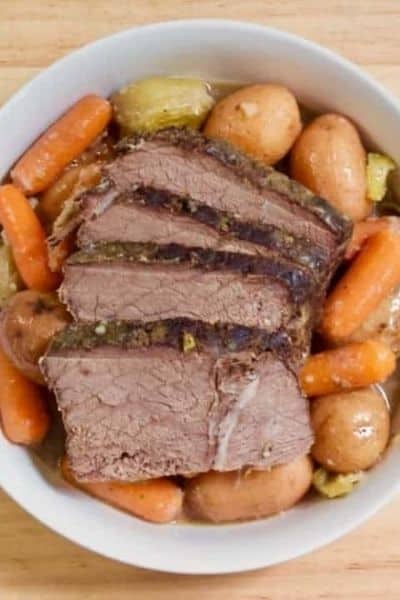 A white bowl on a light wood background filled with mississipi roast including carrots and potatoes