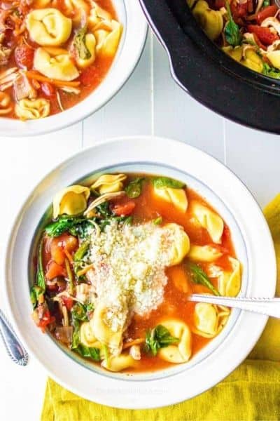 A bowl of slow cooker tortellini soup with a second bowl and the crockpot insert in the background