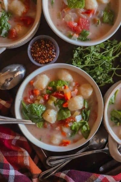 Several bowls of crockpot vegan gnocchi soup with various ingredients in the background