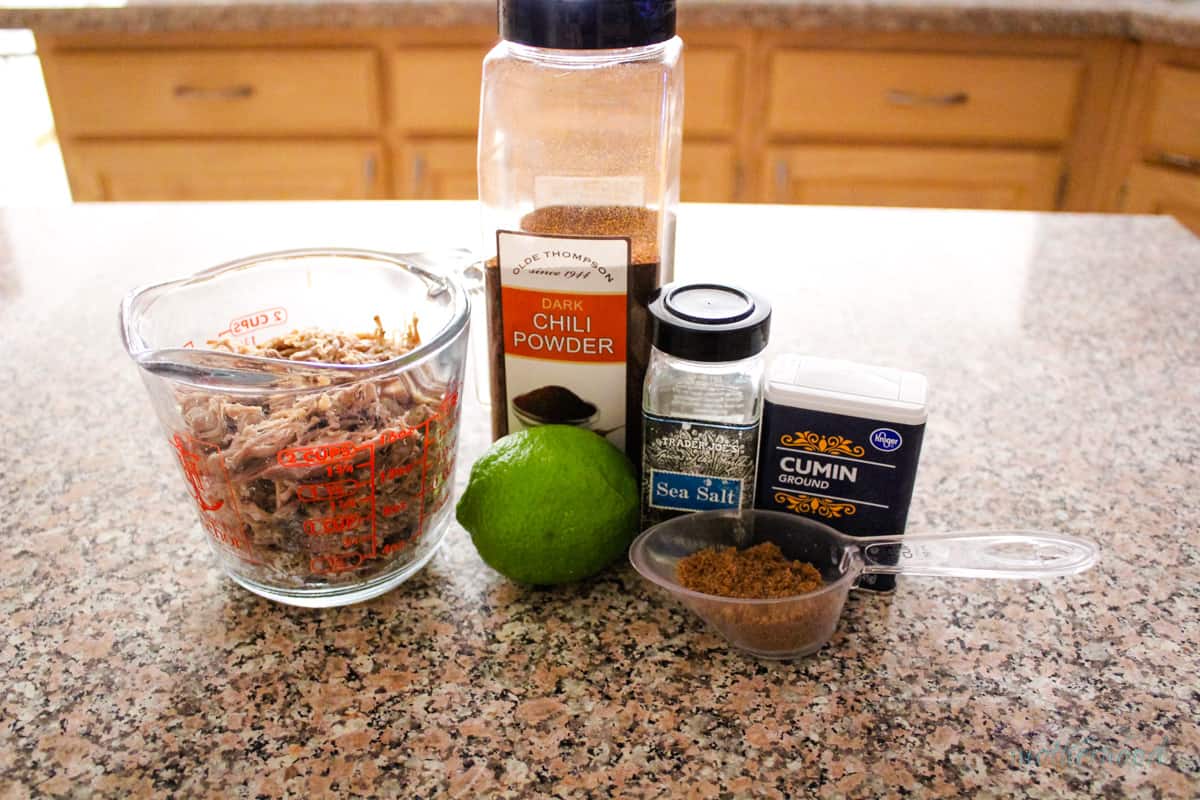 Cooked pulled pork, chili powder, a lime, salt, cumin, and brown sugar on a kitchen counter