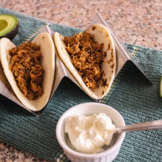 Two tacos in a holder on a counter with avocado, lime, and sour cream surrounding them.