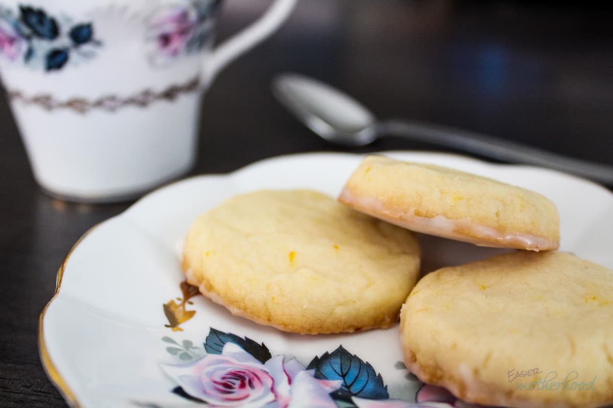 3 cookies on a white tea plate with purple and blue flower painting, a matching teacup in the background along with a spoon.