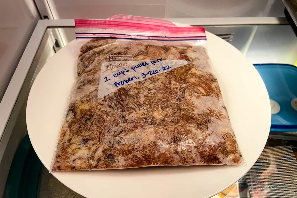 White plate in refrigerator with quart bag of frozen pulled pork sitting on it.