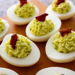 Avocado Deviled Eggs garnished with bacon on a wood serving tray.