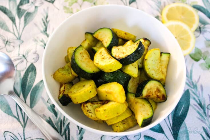 White bowl with zucchini and squash in it on a kitchen towel with lemon slices and a serving spoon.