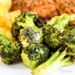 Cheesy broccoli on a white plate with potatoes and chicken in the background