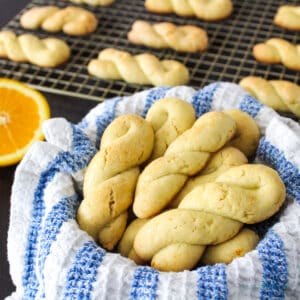 Koulourakia cookies in basket with half an orange and additional cookies on cooling rack in background