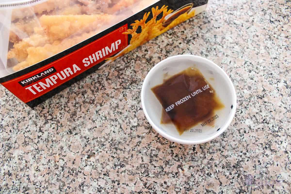 Open box of kirkland tempura shrimp with two packets of sauce in a bowl of water.