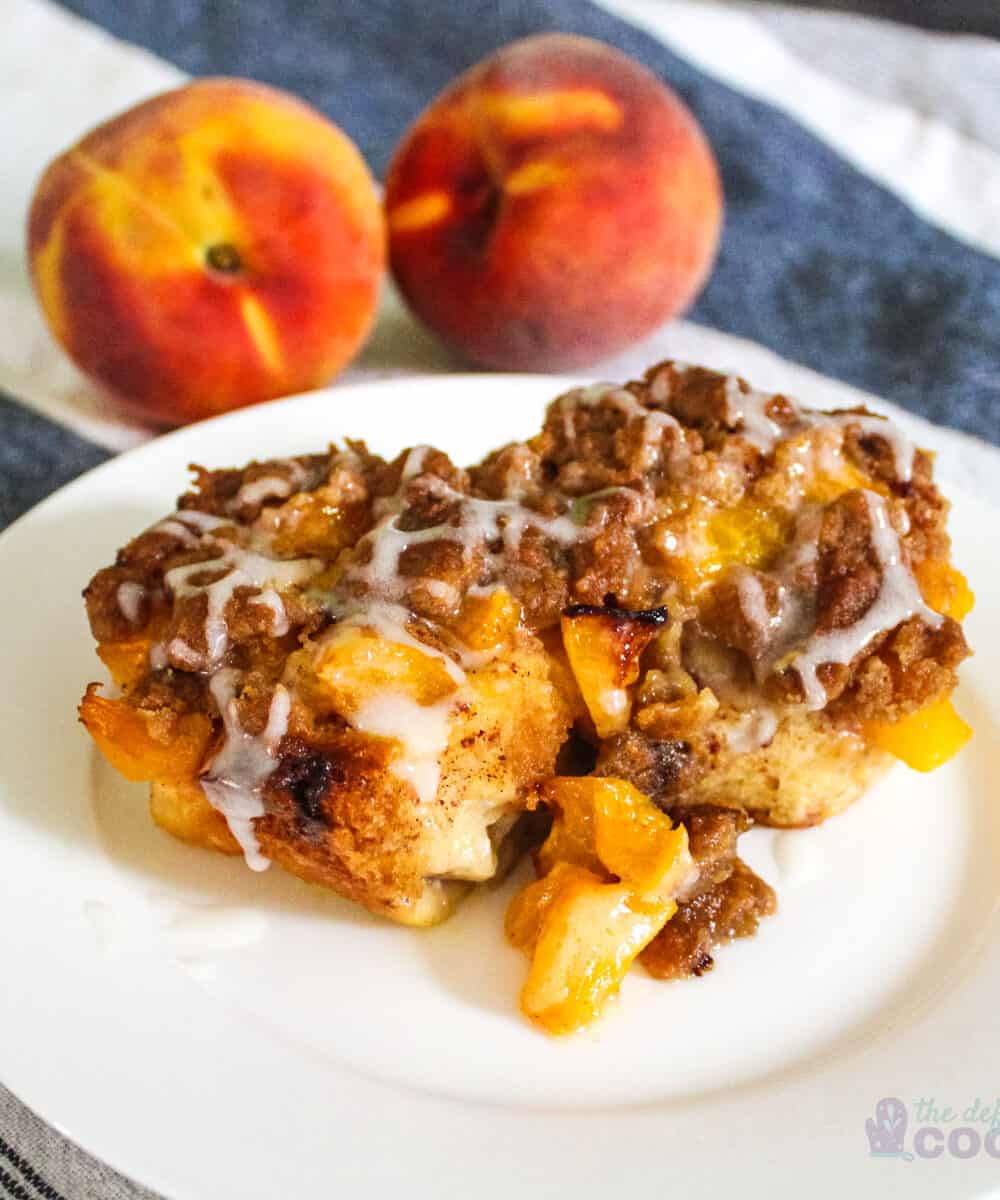 2 peach cobbler cinnamon rolls on a white plate on a kitchen towel with peaches in the background.