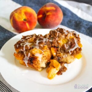 2 peach cobbler cinnamon rolls on a white plate on a kitchen towel with peaches in the background.