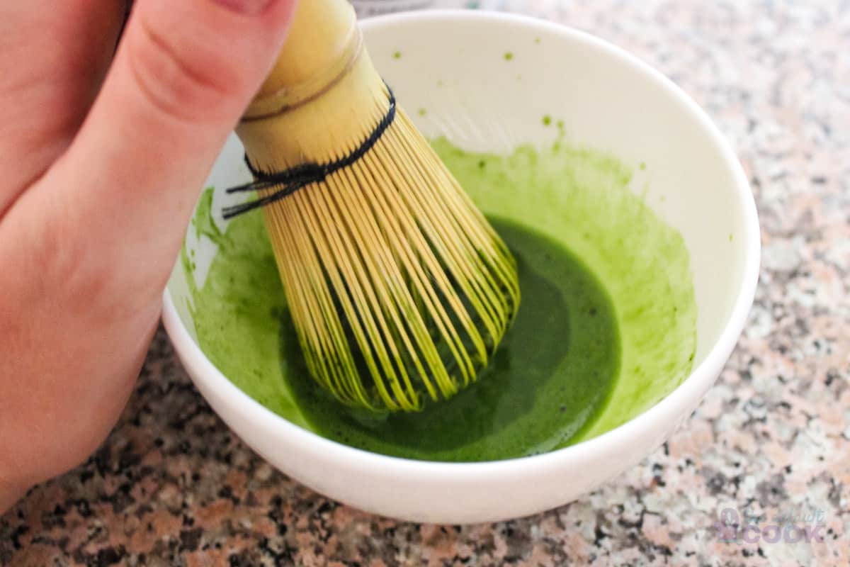 Hand using a bamboo whisk to mix matcha powder and water in white bowl.