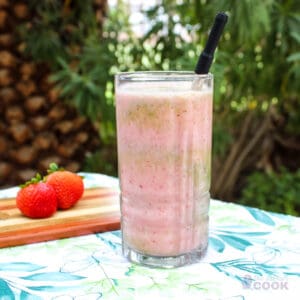Smoothie on a table outdoors with strawberries on a cutting board behind it.
