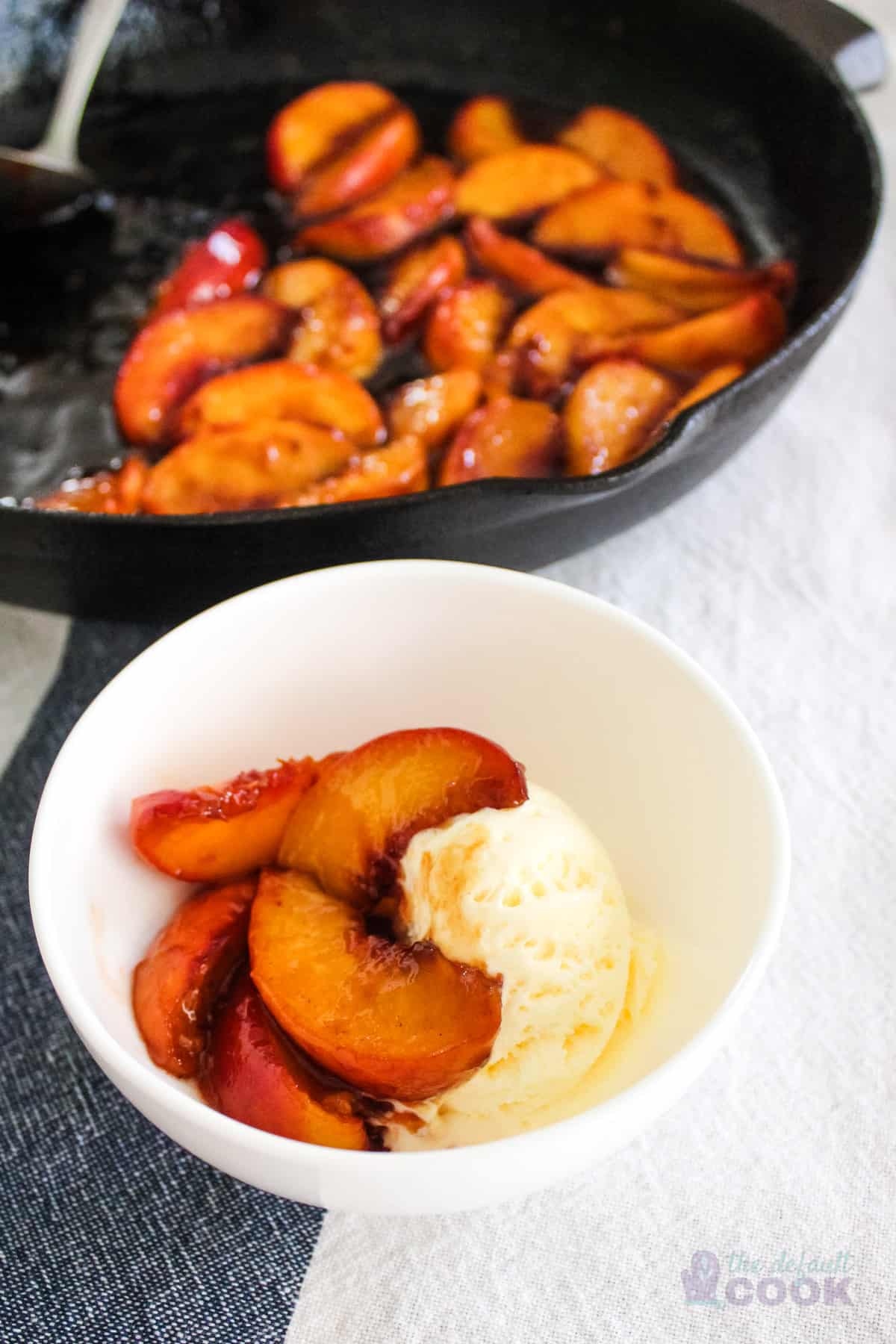 Caramelized peaches on top of vanilla ice cream with skillet of additional cooked peaches in background.