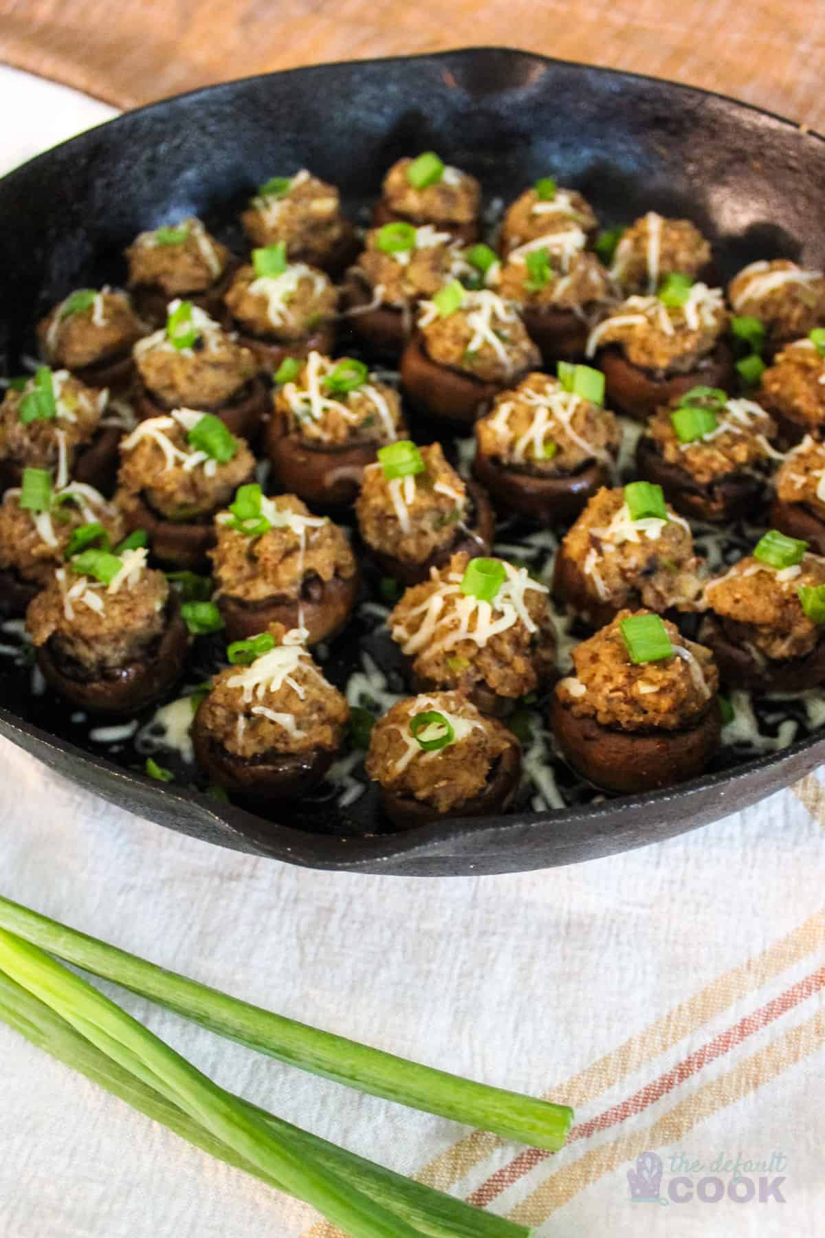 Skillet of stuffed mushrooms on kitchen towel with green onions in front.