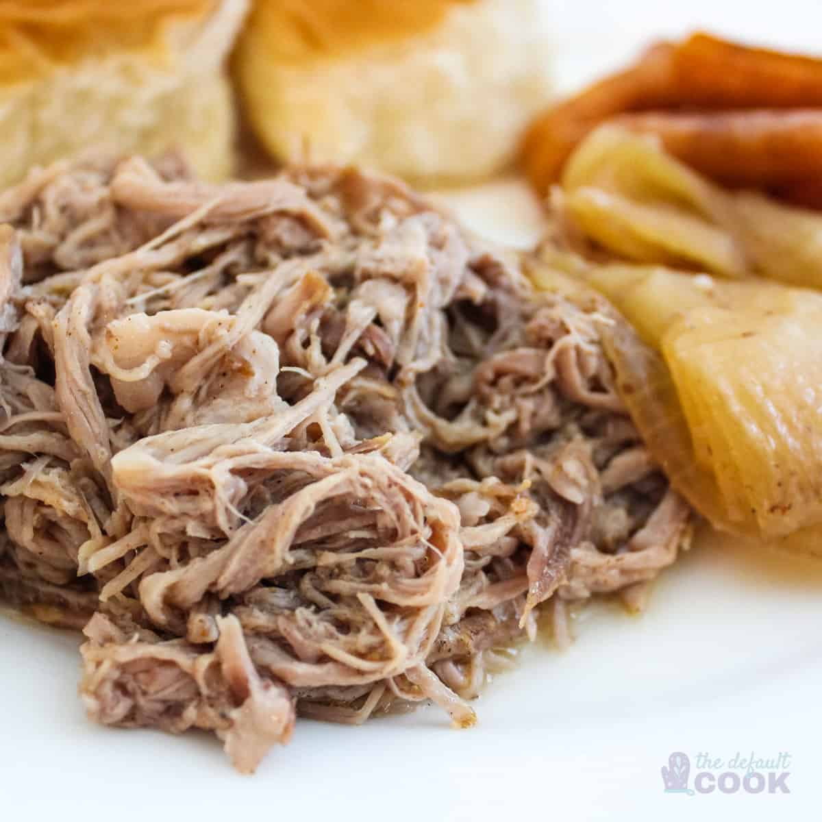 Close up of pulled pork on white plate with onions, carrots, and rolls in background.