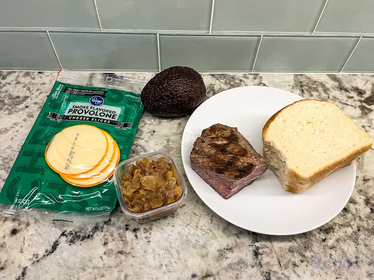 Ingredients on kitchen counter: cheese, caramelized onions, steak, bread, and avocado.