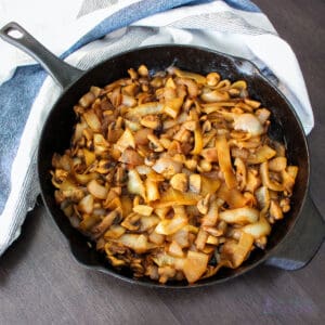 Skillet of caramelized onions and mushrooms on a brown table with blue and white kitchen towel.