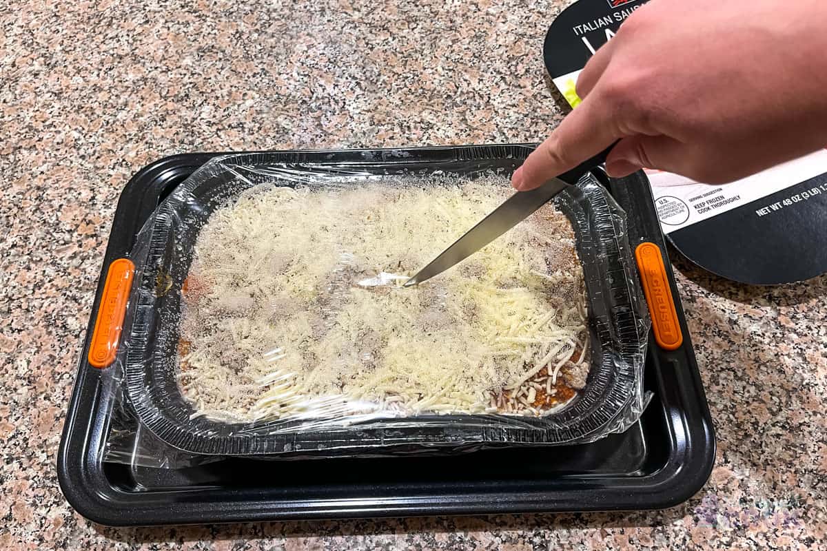 Slicing vent in plastic film of lasagna tray which is on a quarter sheet baking tray.