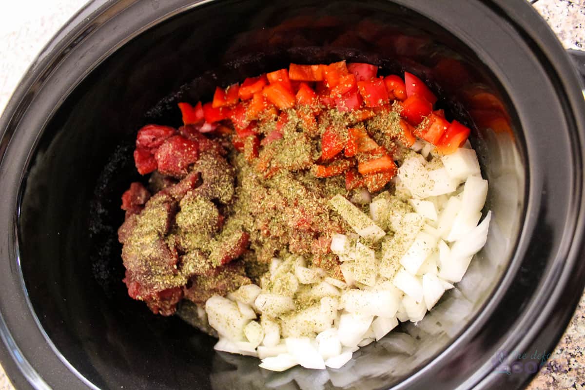 Ingredients in slow cooker insert before stirring, veggies and meat on bottom, seasonings on top.