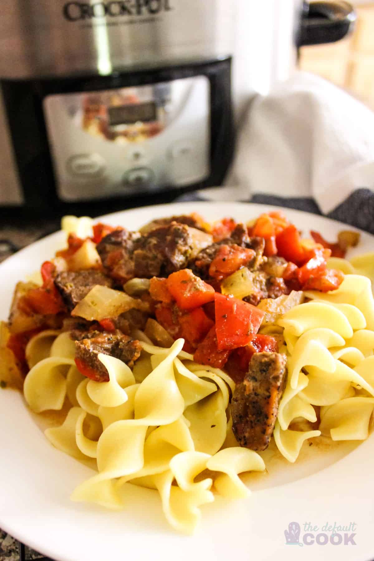 White plate with slow cooker Italian pepper steak on a bed of egg noodles in front of crock pot.