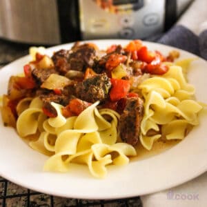 White plate with slow cooker Italian pepper steak on a bed of egg noodles in front of crock pot.