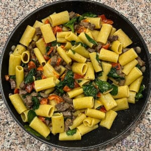 Skillet with tri tip rigatoni pasta with spinach and red peppers on a kitchen counter.