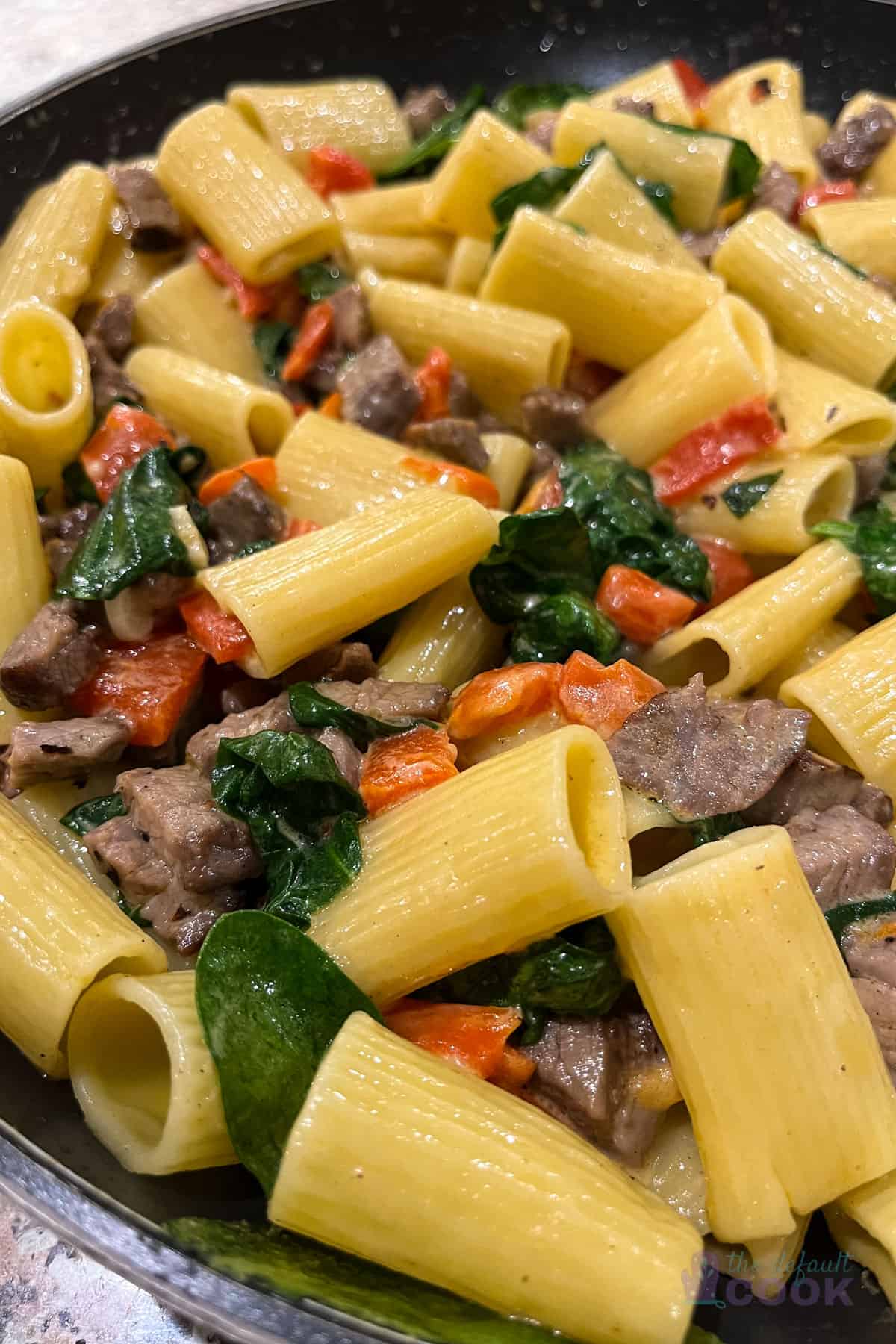 Close up of a skillet of tri tip pasta with red peppers and spinach in a lightly creamy sauce.