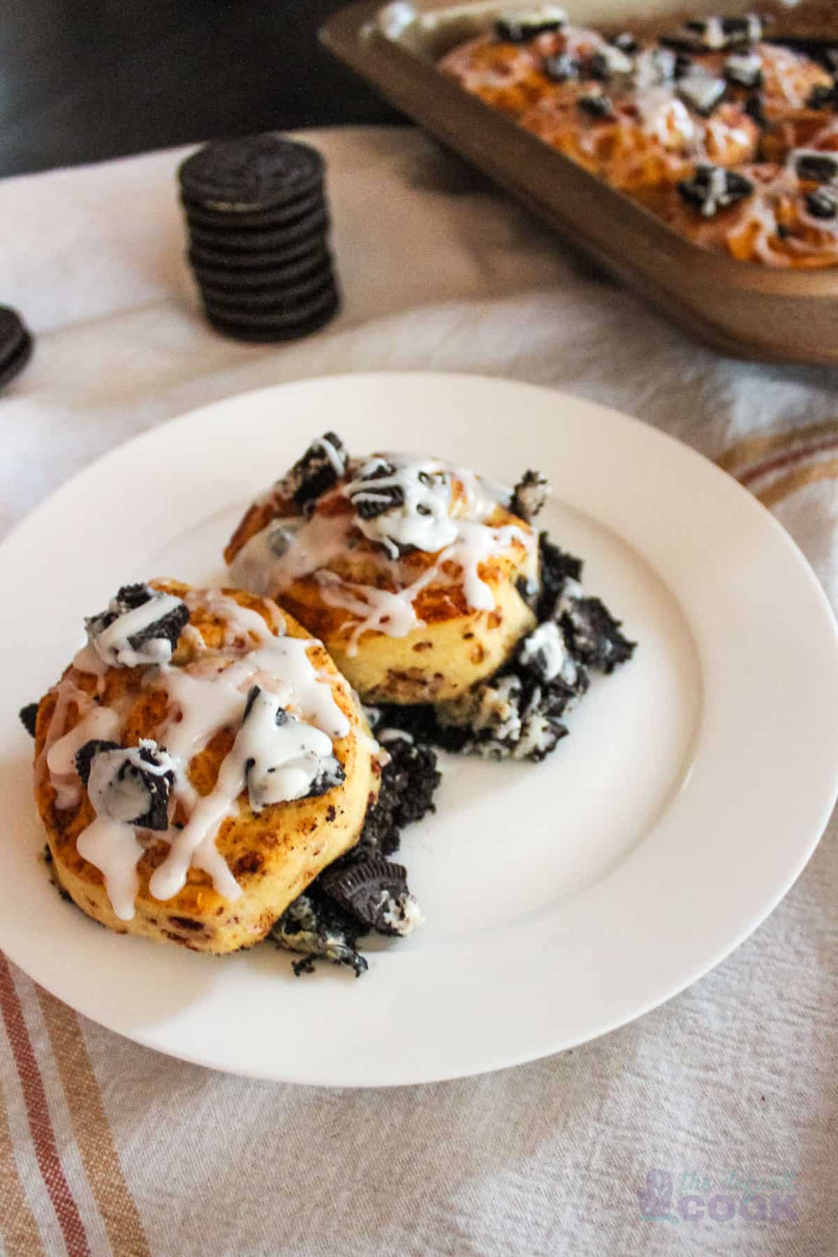 Two oreo cinnamon rolls on a white plate with baking pan and additional oreos in background.