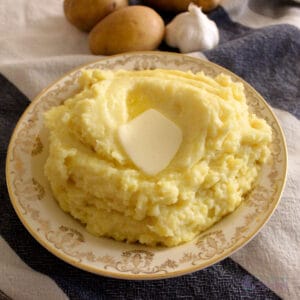 A plate of air fried mashed potatoes with butter melting on top, and raw potatoes and garlic in the background.