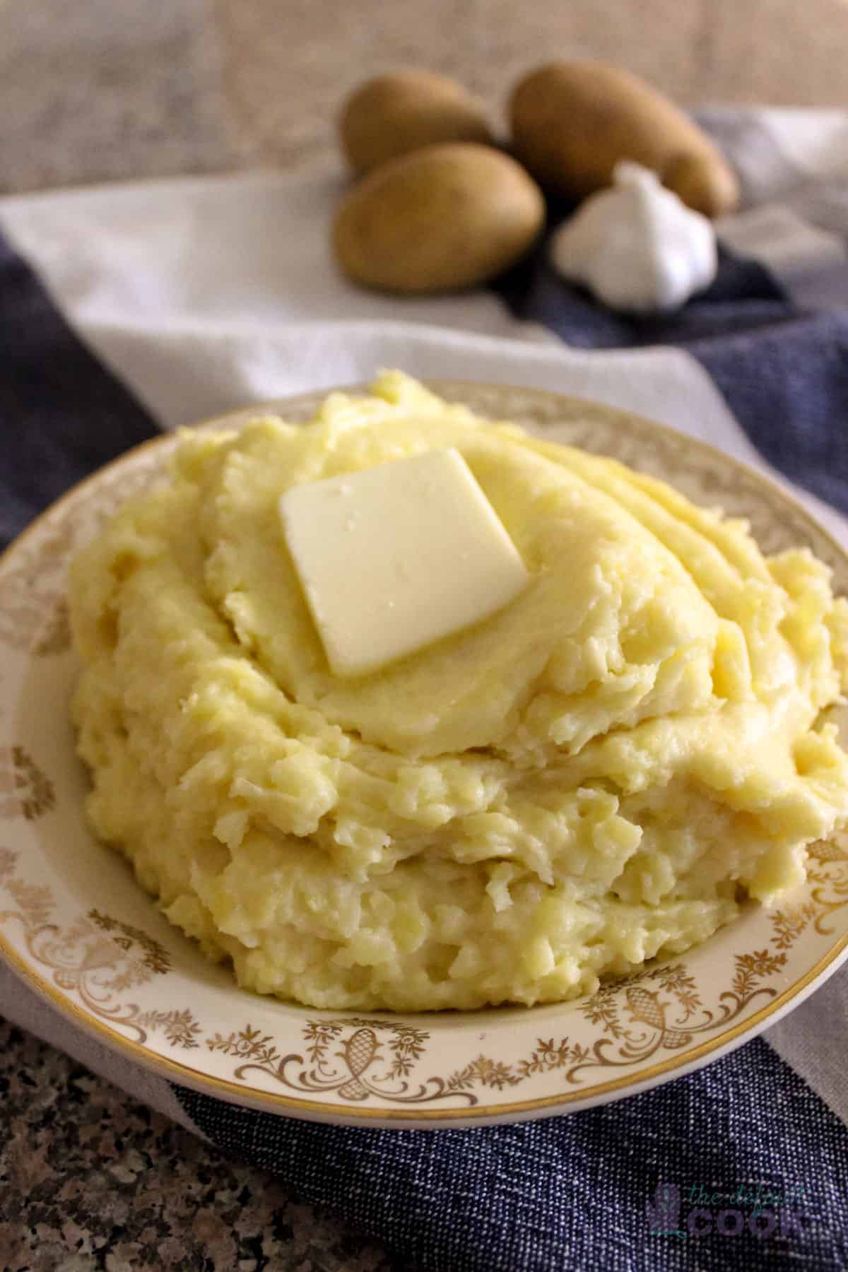 Plate of air fried mashed potatoes with a pat of butter melting on top, potatoes and garlic in the background.