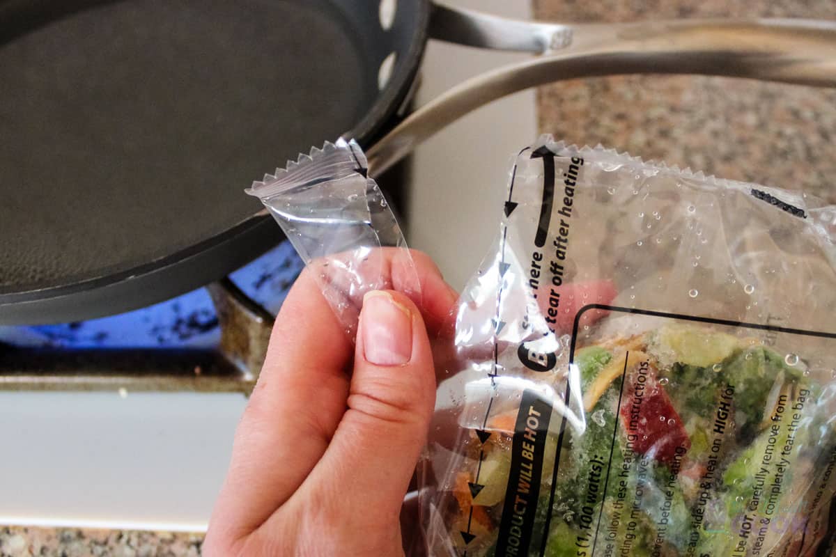 Hand tearing open a package of frozen noodles with a skillet of boiling water in the background.