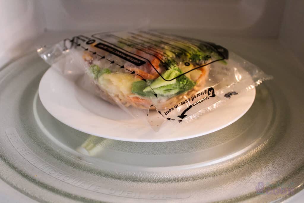 Uncooked frozen yakisoba noodles with vent piece torn, sitting on a white plate in microwave ready to cook.