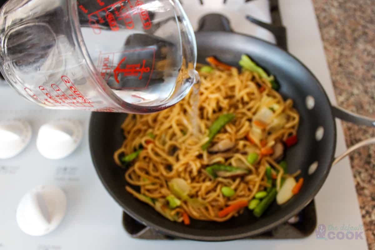 Nearly cooked noodles in skillet with pyrex cup pouring in additional water.