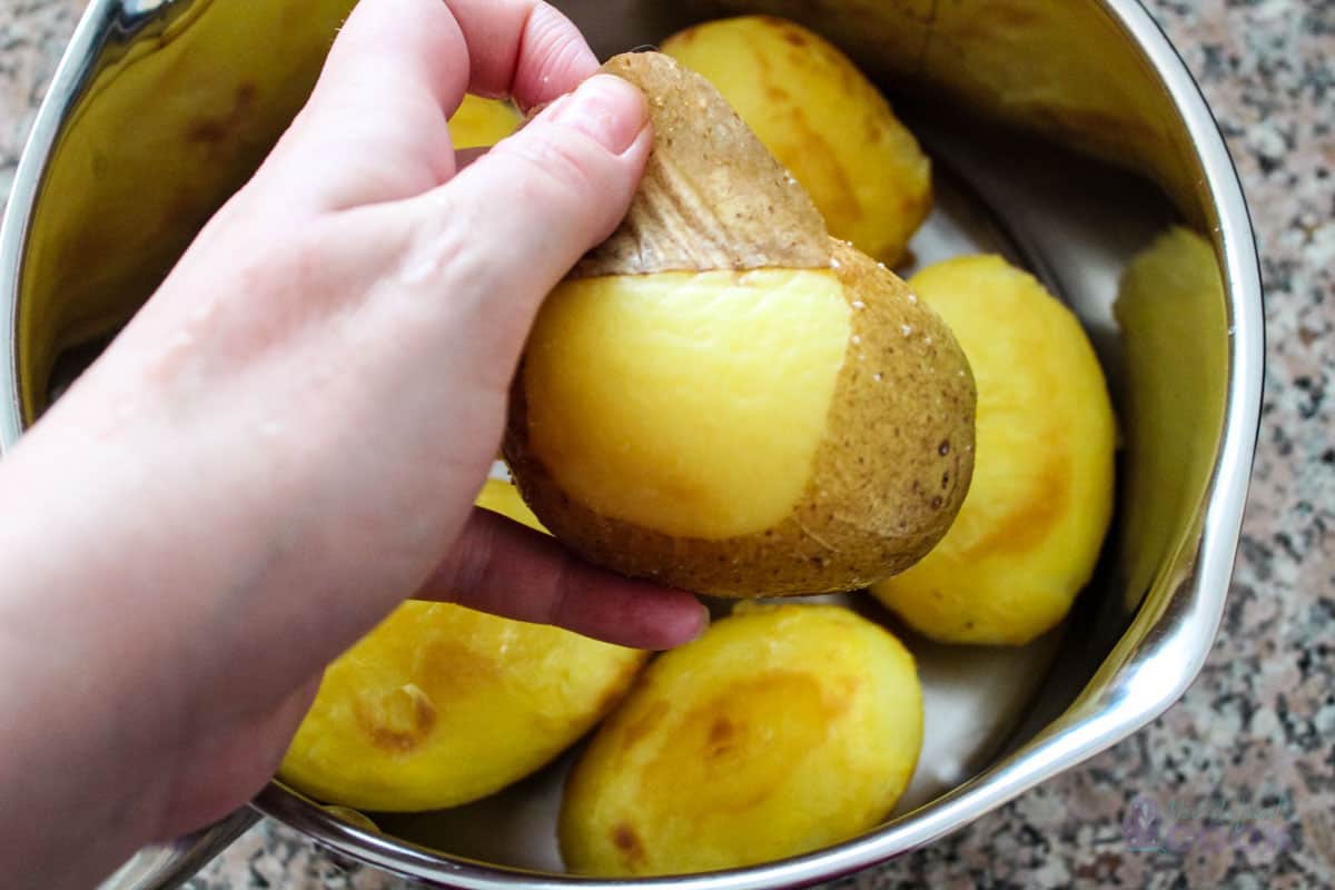 Hand peeling a potato with a pan of the other peeled potatoes in the background.