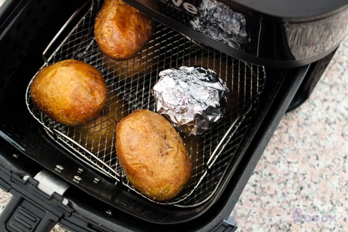 Cooked potatoes and head of garlic wrapped in aluminum foil in a basket style air fryer.