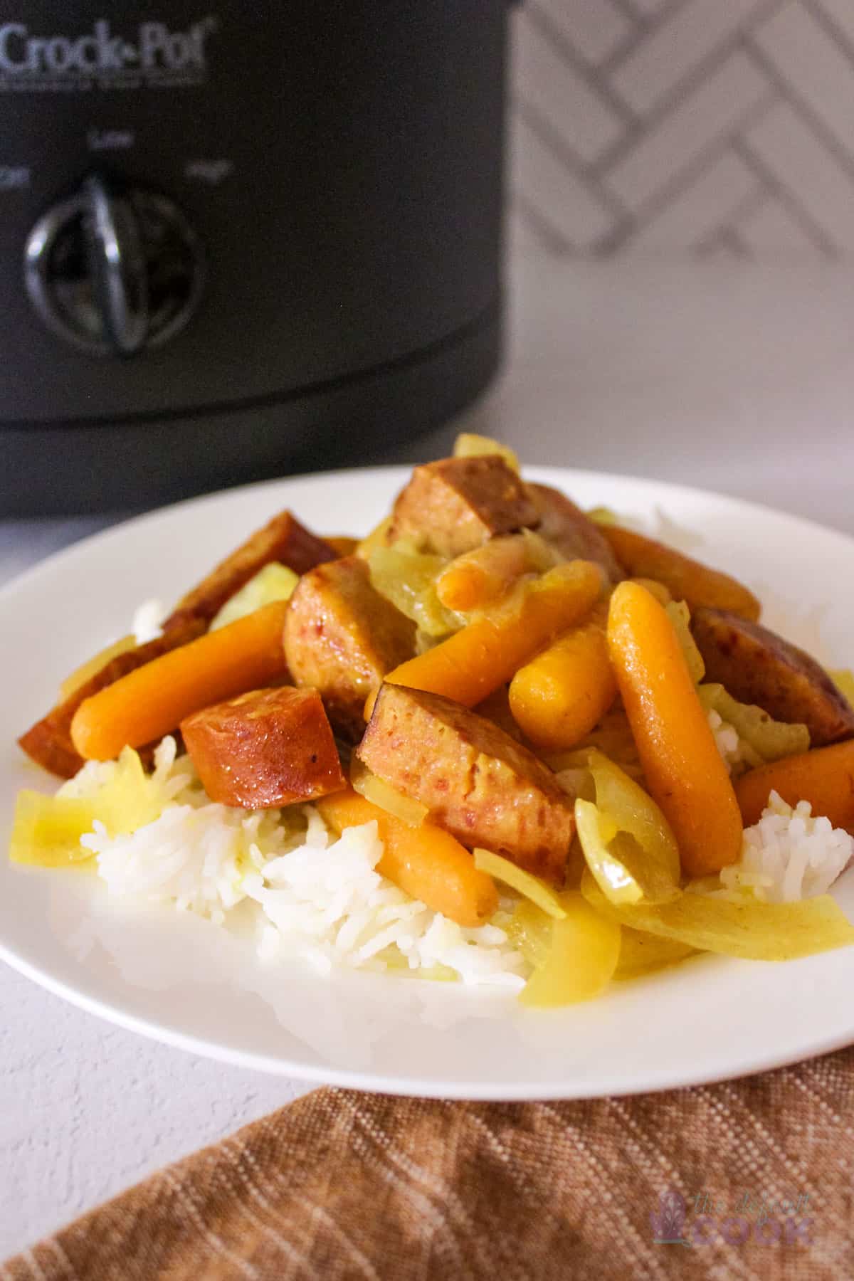 Plate of coconut curry sausage and vegetables on a bed of rice in front of a slow cooker.