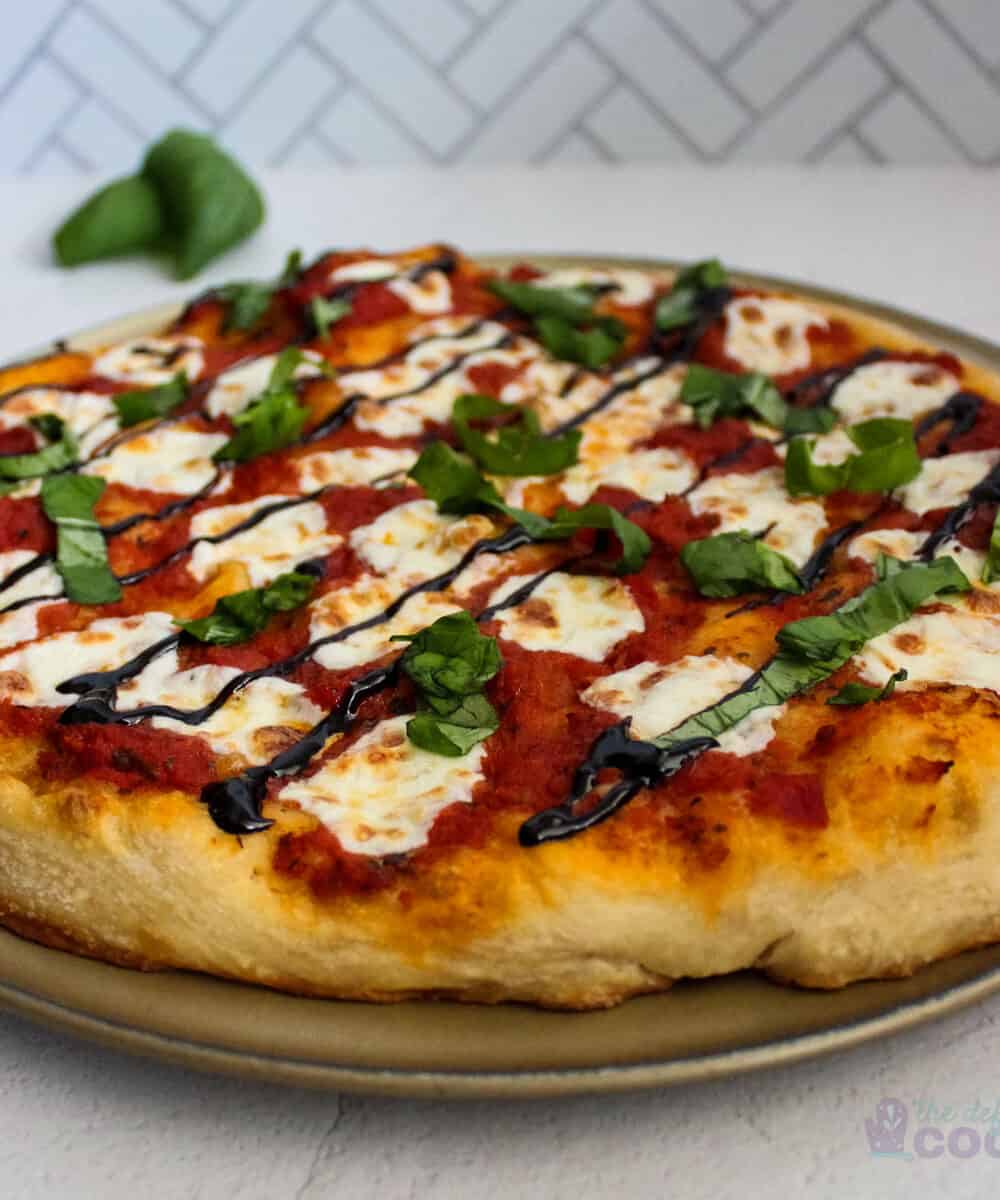 Pizza on the pan with basil behind it, both on a white marble surface with chevron tile backsplash in the background.
