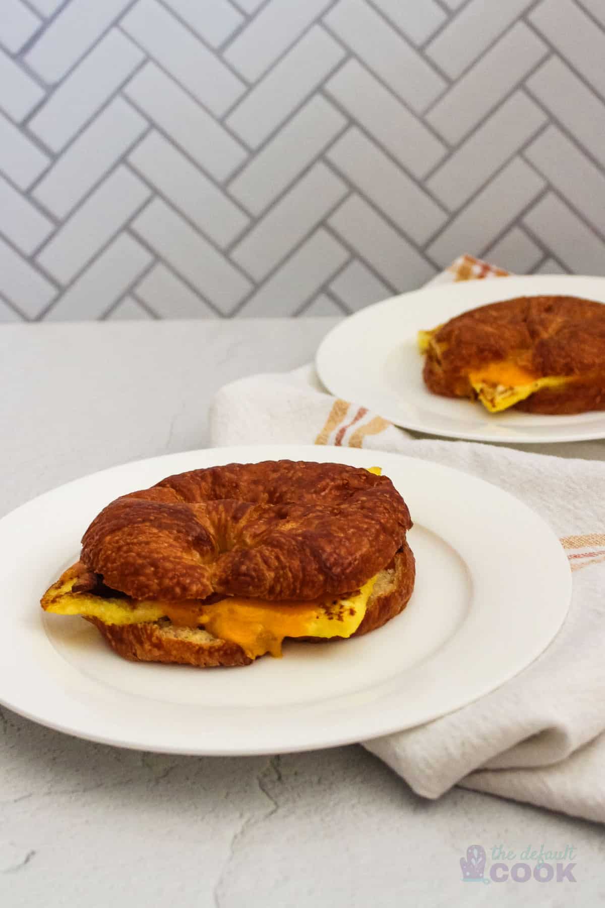 Two breakfast croissant sandwiches on white plates on a counter.