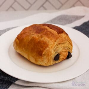 Chocolate croissant on a white plate with a blue and white kitchen towel under it.