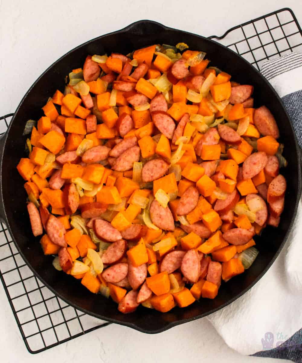 Overhead of an iron skillet with cooked sweet potatoes, onions, and sausage on a wire cooling rack with a kitchen towel under part of it.