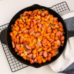 Overhead of an iron skillet with cooked sweet potatoes, onions, and sausage on a wire cooling rack with a kitchen towel under part of it.