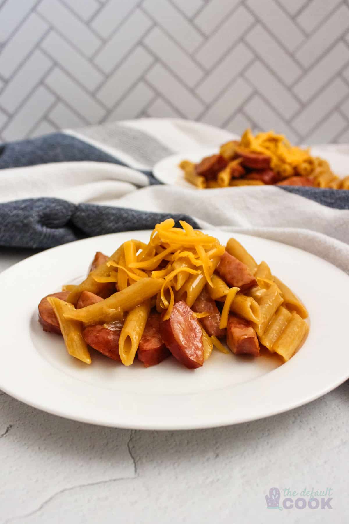 Two plates of cheesy kielbasa pasta on a kitchen counter with a kitchen towel.