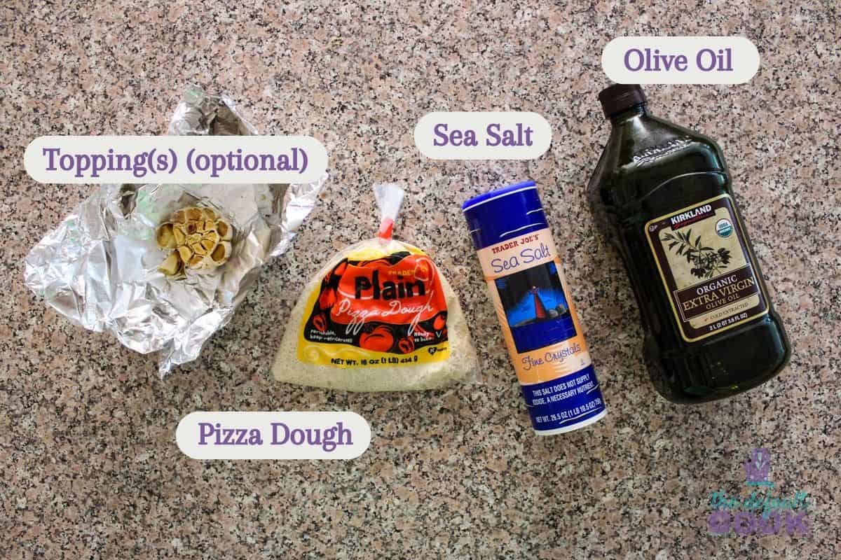 Labeled ingredients sitting on a counter: roasted garlic, pizza dough, sea salt, and olive oil.
