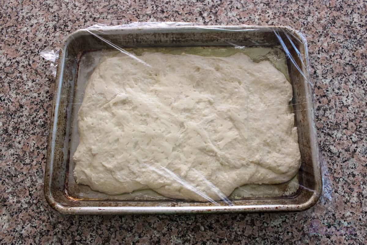 Pizza dough on quarter sheet pan with plastic wrap over it.